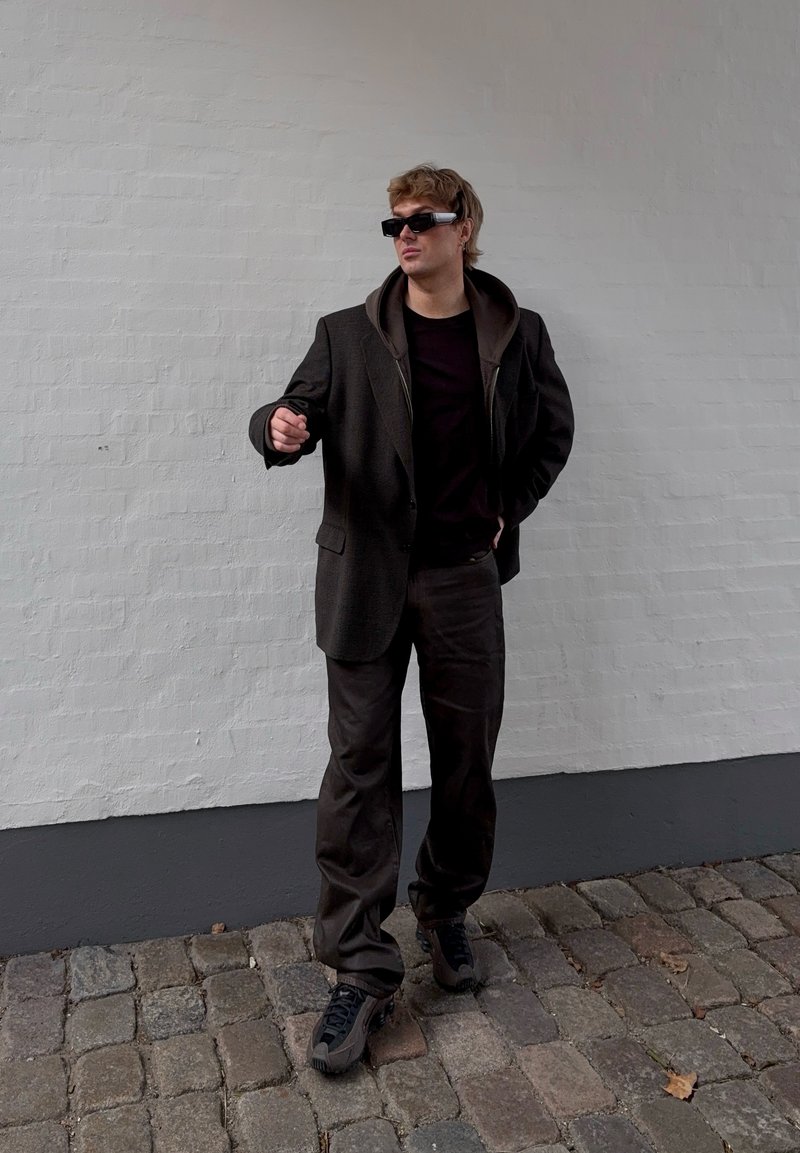 Moški nosi črn prevelik blazer, kapucar, temne hlače, debele superge in futuristična črna sončna očala, stoji na tlakovani ulici.
