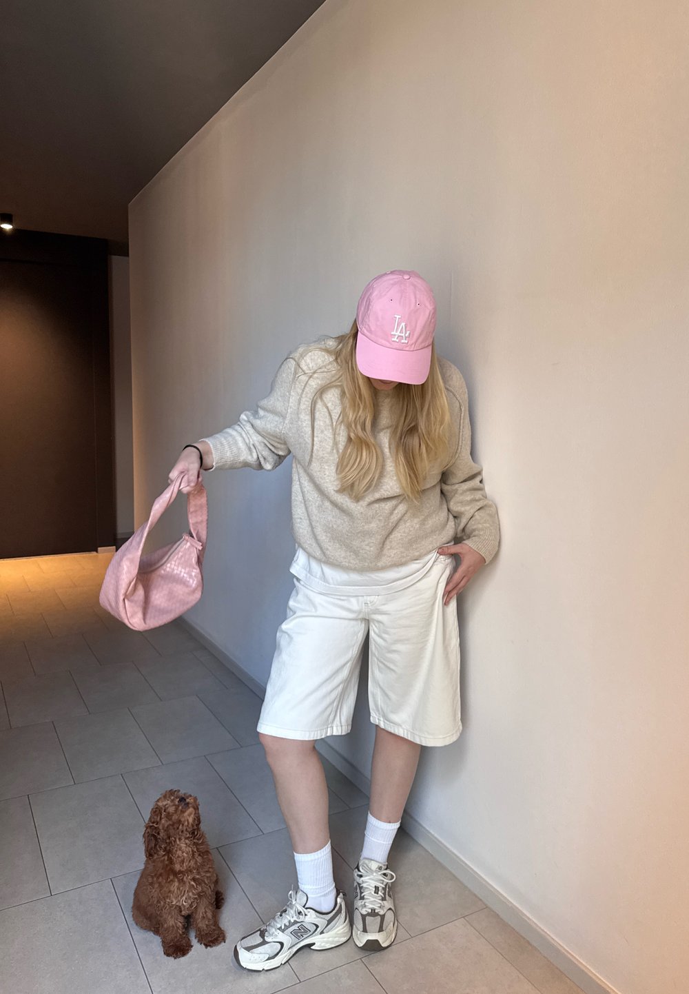 Hellgrauer Strickpullover, weiße Shorts, rosa Kappe und rosa strukturierte Tasche. Weiße SportSneaker und Knöchelsocken. Kleiner brauner Hund sitzt in der Nähe.