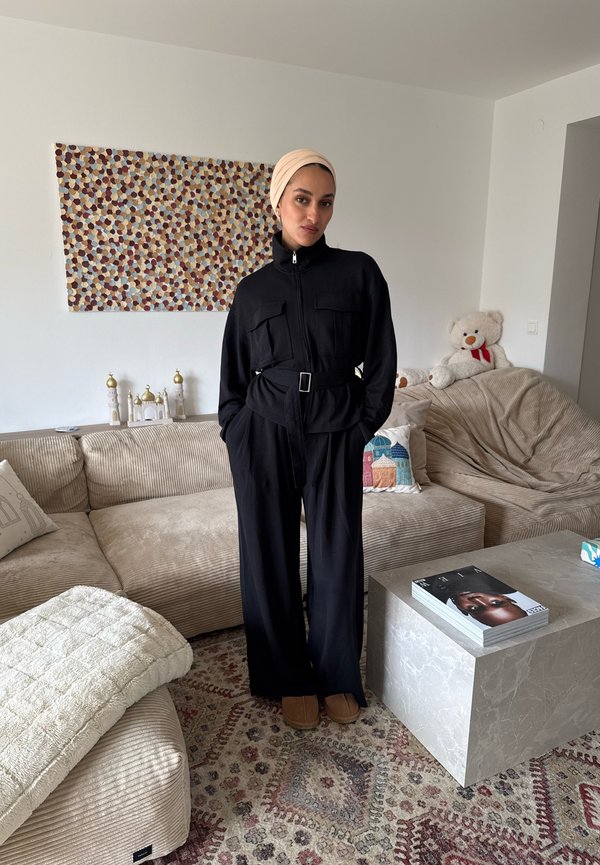 Mujer con chaqueta negra con cinturón, pantalones holgados, pañuelo beige en la cabeza y zapatillas, de pie en una sala de estar acogedora con alfombra estampada y oso de peluche en el sofá.