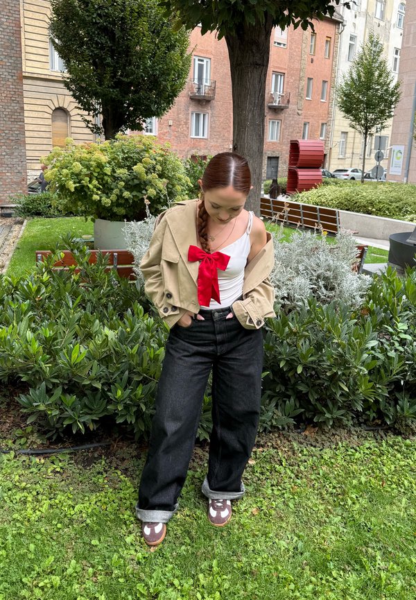 Joven mujer con cabello trenzado, vistiendo chaqueta beige, top blanco con lazo rojo, pantalones negros, de pie sobre el césped en un jardín urbano con arbustos y edificios.