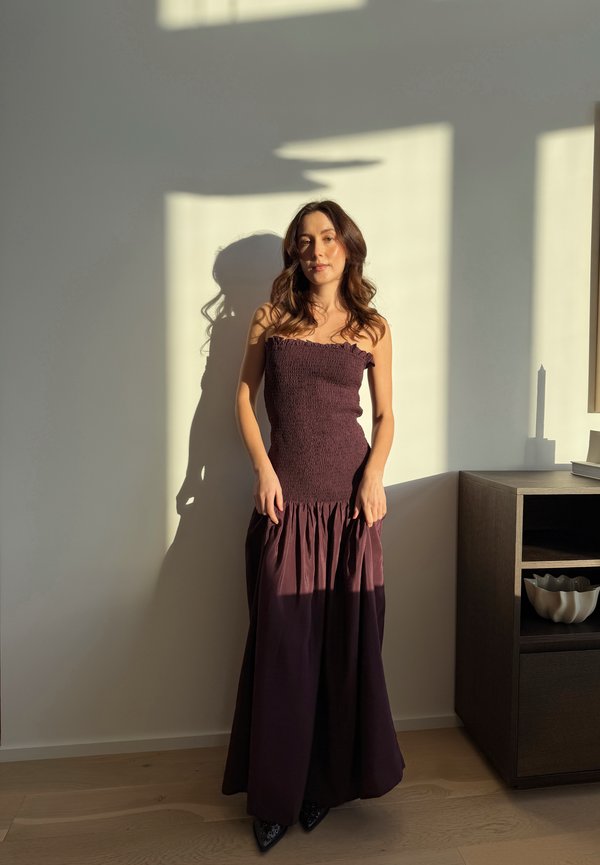 Fiatal nő ujjatlan sötétlila ruhában, beltéren áll, a napfény árnyékokat vet a falra és a fapadlóra egy polcos szekrény mellett.