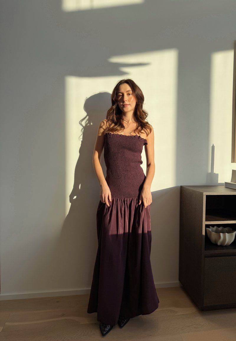 Jeune femme en robe sans manches couleur violet foncé, debout à l'intérieur, la lumière du soleil projetant des ombres sur le mur et le sol en bois à côté d'une étagère.