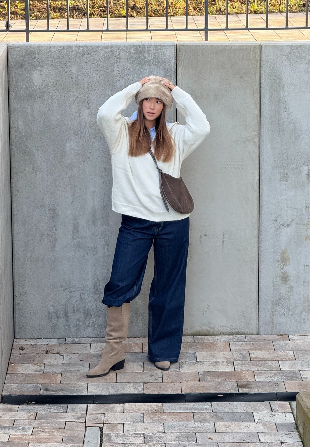 Fehér kötött pulóver, sötétkék bő szárú farmer, bézs térdig érő csizma és világosbarna szőrmekalap, beton háttér előtt állva.