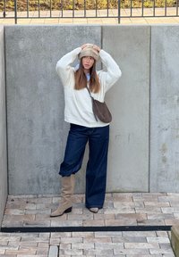 Fehér kötött pulóver, sötétkék bő szárú farmer, bézs térdig érő csizma és világosbarna szőrmekalap, beton háttér előtt állva.