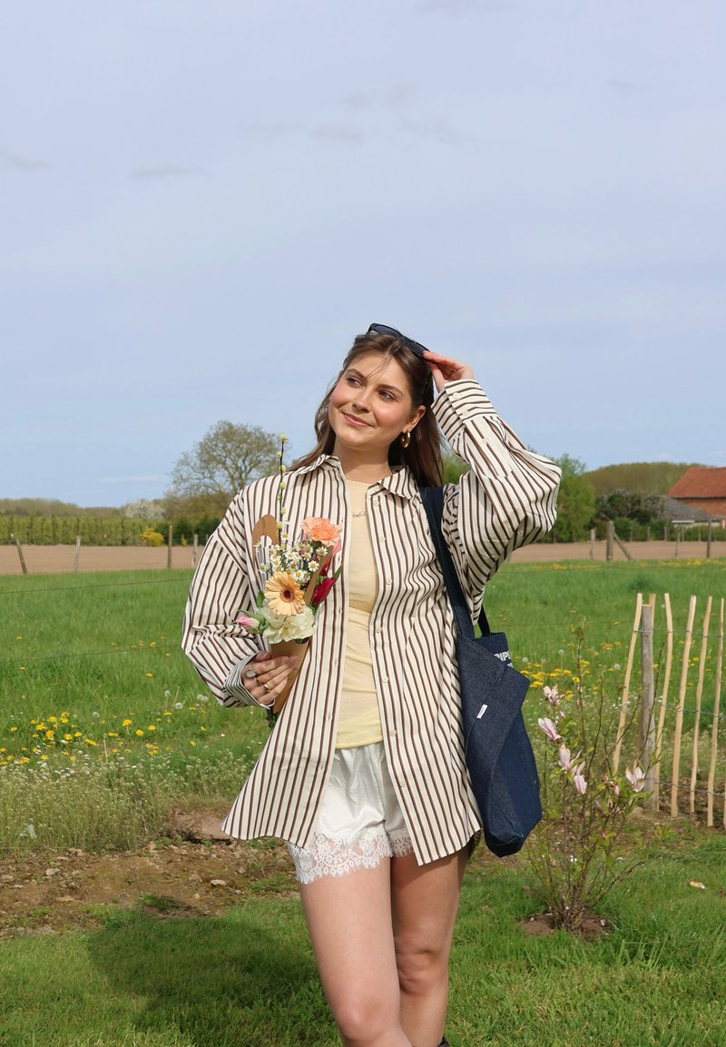 Jeune femme en chemise à rayures et short tient un bouquet, debout dans un champ herbeux avec des fleurs et des arbres en arrière-plan.
