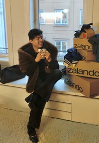 Brun kunstpels-wrap, sort top, mørke bukser og sko med hæl. Sidder på en hvid overflade ved siden af Zalando-kasser og holder en hvid krus.