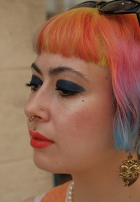 Close-up van een persoon met multicolore haar (oranje, roze, geel), donkere oogschaduw, oranje lippenstift en een grote hartvormige oorbel.