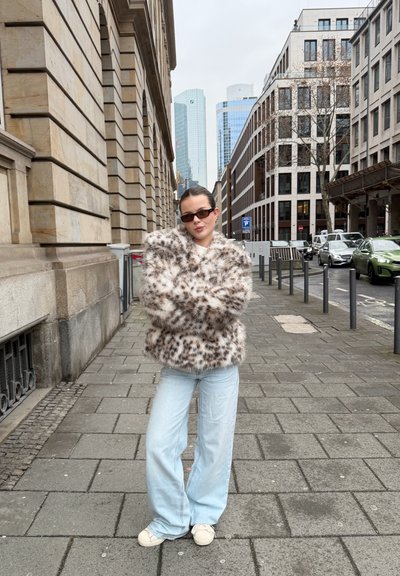 Joven con gafas de sol que lleva una chaqueta de piel sintética con manchas, jeans azul claro y zapatillas blancas, de pie en una acera de la ciudad.