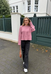 Różowy dzianinowy sweter z długimi rękawami, zestawiony z czarnymi spodniami o szerokich nogawkach i białymi trampkami. Trzyma małą brązową torebkę. Tło z kamiennej kostki.