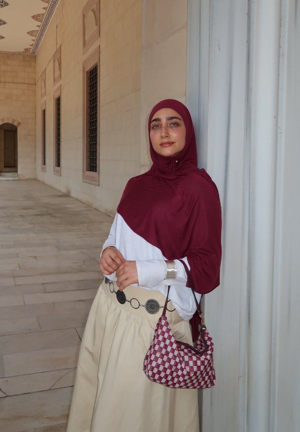 Maroon huivi ja paita valkoisilla hihoilla, beige hame, hopeinen vyö pyöreillä yksityiskohdilla ja ruutukuvioinen maroon laukku. Kiviseinätausta.