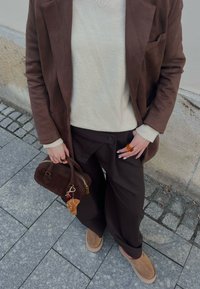 Personne portant un blazer marron, un pull crème, un pantalon large foncé et des chaussures couleur havane, tenant un sac à main marron avec un porte-clés pâtisserie en peluche sur un trottoir pavé.