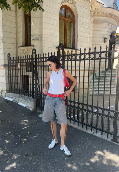 Top blanco sin mangas, cinturón rojo, pantalones cortos de mezclilla desgastados, zapatillas blancas con detalles negros, gafas de sol reflectantes, de pie junto a una valla de hierro forjado.