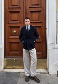 Jūrų laivyno dvigubas paltas ant šviesiai mėlynos marškinėlės ir rudos kaklaraiščio, derinamas su šviesiai bežiais kelnėmis ir rudomis bateliais, stovintis prieš medines duris.