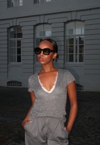 Femme portant des lunettes de soleil noires, un haut en maille gris avec bordure en dentelle et un pantalon gris à rayures, debout les mains dans les poches devant une façade de bâtiment.