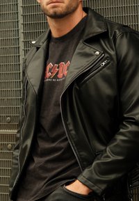 Jaqueta de motoqueiro em pele sintética preta com zíperes prateados, sobre uma t-shirt gráfica escura. Textura suave e design ajustado.