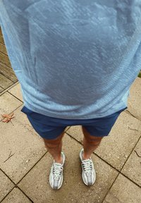 Camisola desportiva texturizada azul clara, calções azul marinho e sapatilhas de corrida cinzentas com detalhes verdes, de pé numa calçada de pedra.