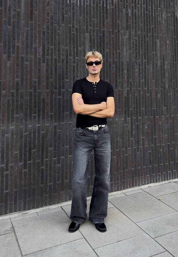 Giovane uomo con capelli biondi e occhiali da sole in piedi con le braccia incrociate, indossa una camicia nera, jeans grigi, cintura bianca e scarpe nere davanti a un muro di mattoni scuro.