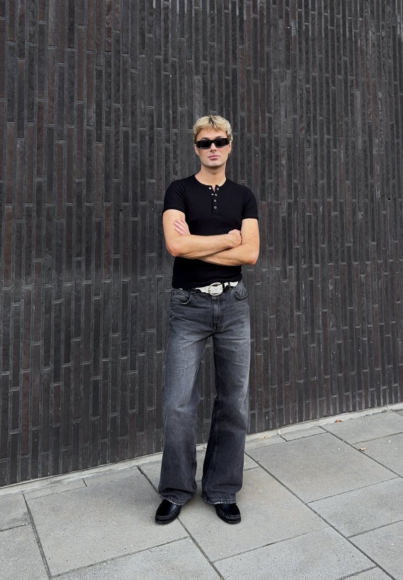 Ung man med blont hår och solglasögon står med armarna i kors, klädd i svart skjorta, grå jeans, vitt bälte och svarta skor framför en mörk tegelvägg.