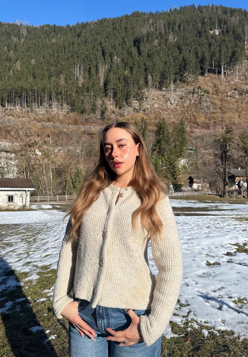Ung kvinne iført beige cardigan og jeans står utendørs med snøflekker, skogkledd ås og klar blå himmel i bakgrunnen.