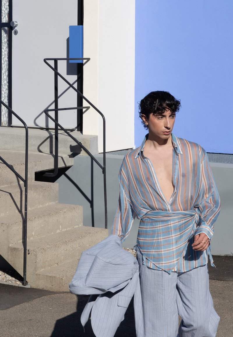 Jeune personne aux cheveux bouclés foncés, portant une chemise ample rayée et un pantalon, tenant une veste légère, debout près d'escaliers en béton et d'un mur bleu.
