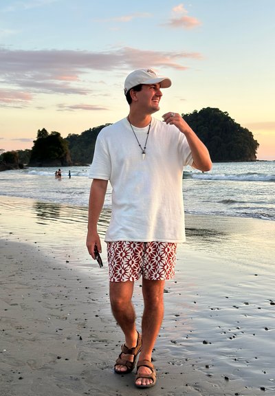 Hombre con gorra blanca, camisa blanca y pantalones cortos estampados caminando descalzo por la playa mojada al atardecer, sosteniendo gafas de sol y sonriendo.