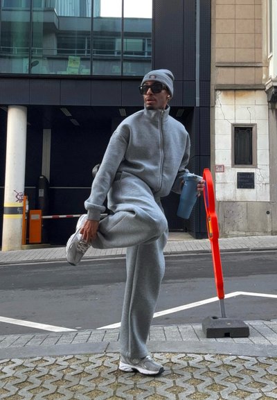 Hombre con chándal gris, gorro y gafas de sol estira la pierna mientras sostiene una botella mezcladora azul en una calle urbana.
