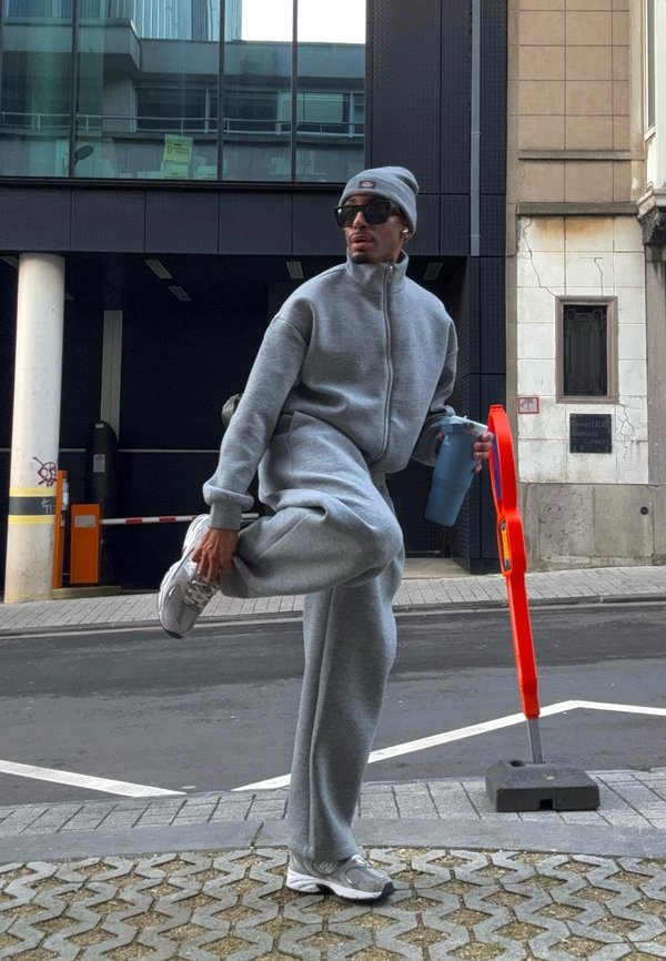 Mężczyzna ubrany w szary dres, czapkę i okulary przeciwsłoneczne rozciąga nogę, trzymając niebieską butelkę shakera na miejskiej ulicy.