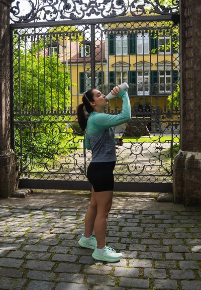 Una mujer con una camiseta de manga larga en tonos verde azulado degradé y pantalones cortos negros bebe de una botella de agua blanca frente a una puerta decorativa de hierro forjado.
