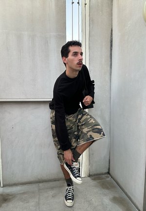 Hombre con camisa negra de manga larga y pantalones cortos de camuflaje se agacha para atarse unas zapatillas negras de caña alta en una estrecha escalera de hormigón.