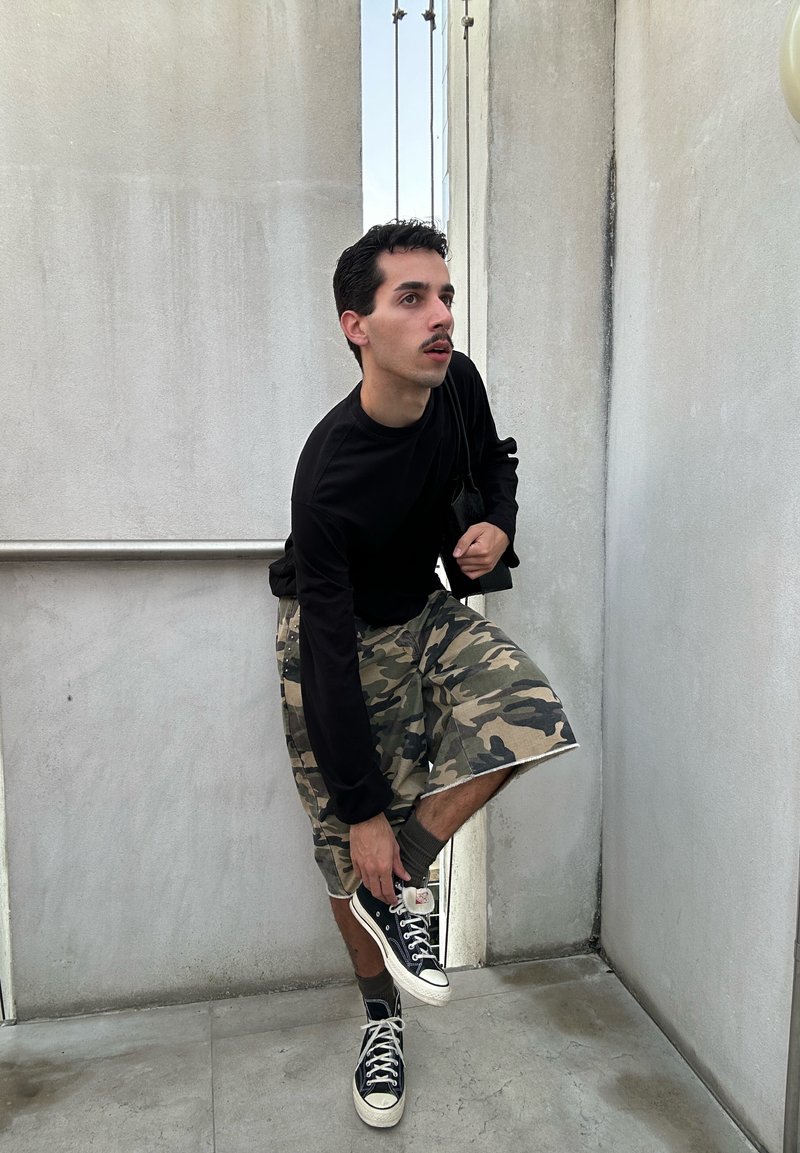 Άνδρας με μαύρο μακρυμάνικο πουκάμισο και κοντά καμουφλάζ παντελόνια σκύβει για να δέσει μαύρα ψηλά αθλητικά παπούτσια σε ένα στενό σκυρόδεμα σκαλοπάτι.