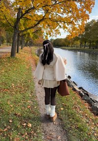 Crème kleurige jas met wijde mouwen, korte geplooide rok, donkere maillots, witte kniekousen en bruine plateauschoenen; draagt een bruine tas.