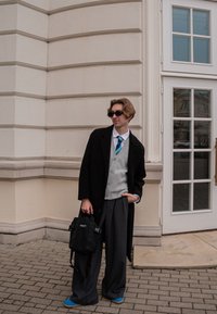 Manteau noir, gilet gris, chemise blanche, cravate bleue, pantalon gris ample, sac noir et chaussures bleues. Debout contre un mur de bâtiment.