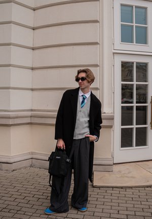 Abrigo negro, chaleco de lana gris, camisa blanca, corbata azul, pantalones grises holgados, bolso negro y zapatos azules. De pie junto a una pared de un edificio.