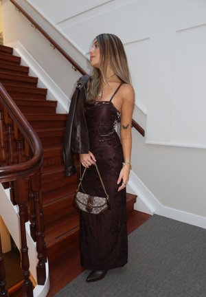 Mujer con un vestido largo floral oscuro y botas, de pie cerca de una escalera de madera, sosteniendo un bolso estampado y una chaqueta de cuero sobre un hombro.