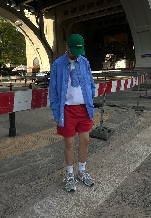 Joven con gorra verde, camisa azul, camiseta blanca con diseño, pantalones cortos rojos, calcetines blancos y zapatillas grises, de pie en una calle de la ciudad cerca de barreras.