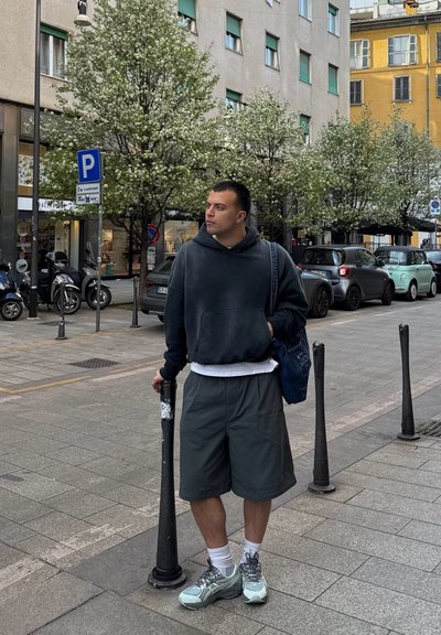 Conjunto informal que incluye una sudadera oscura, una camiseta blanca, pantalones cortos grises y zapatillas grises. Entorno urbano con árboles en flor, coches aparcados y señales de tráfico.