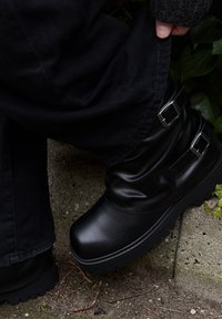 Juodos odos kulkšnies batai su laisvos formos dizainu, dviem sidabriniais sagtimis ir storu juodu guminiu padu. Viršus yra lygios tekstūros.