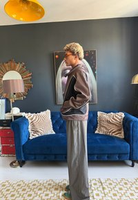 Kiharat blondit hiukset ja silmälasit omaava henkilö seisoo sivuttain olohuoneessa, jossa on sininen sohva, kuvioidut tyynyt ja taidetta seinällä takana.