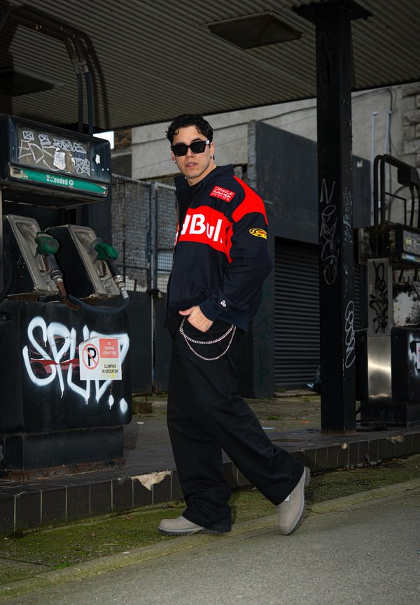 Mann mit Sonnenbrille und schwarzer-rote Jacke steht neben mit Graffiti bedeckten, alten Zapfsäulen an einer verlassenen Tankstelle.