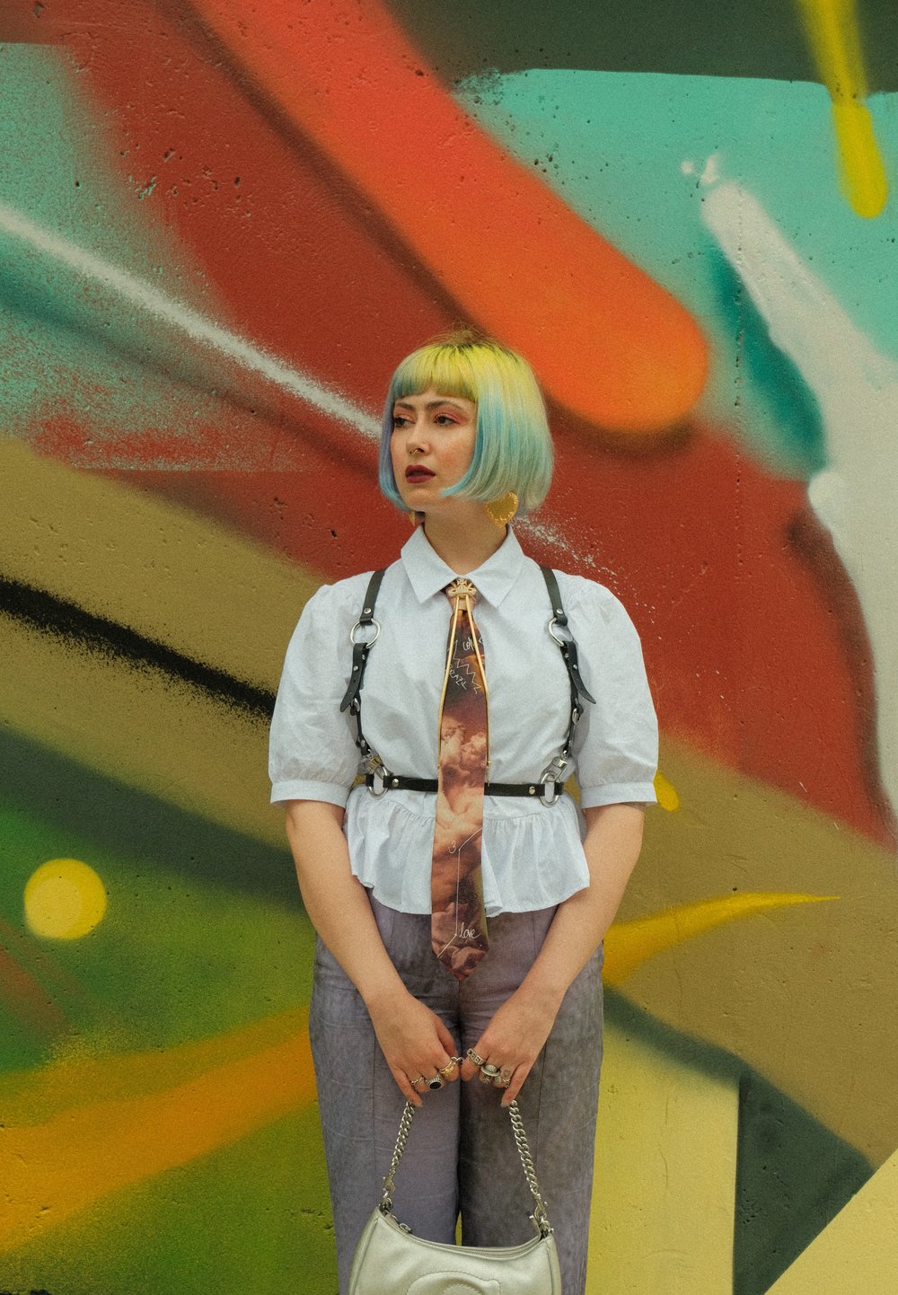 Hvid kortærmet bluse med flæsekant, sort sele, mønstret slips, lyse lilla bukser og sølvfarvet håndtaske foran en farverig graffiti-baggrund.
