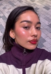 Lähikuva henkilöstä, joka pukeutuu violetti-valkoisiin raidallisiin poolopaitaan. Henkilöllä on kiiltävät punaiset huulet, niittikorvakorut ja sileä, terve iho.