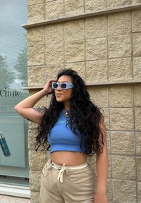 Occhiali da sole azzurri, top corto a coste blu, pantaloni beige a vita alta con nodo. Capelli lunghi e ricci con uno sfondo di muro in pietra testurizzato.