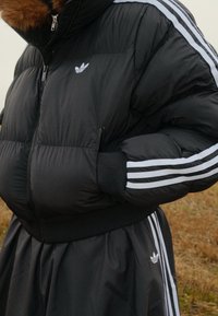 Doudoune noire avec capuche doublée de fourrure, ornée de trois bandes blanches sur les manches et d'une poche avant zippée, associée à une jupe noire.