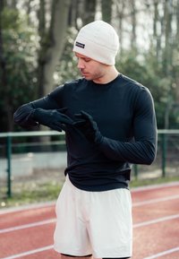 Urheilullinen mies mustassa pitkähihaisessa paidassa ja valkoisissa shortseissa säätää mustia hanskoja, päässään kermaisen vaalea pipo, punaisella kenttäpinnalla.