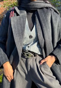 Femme portant un manteau à rayures grises superposé, un cardigan gris clair boutonné, un col roulé noir, un pantalon gris et une ceinture avec une grande boucle argentée.