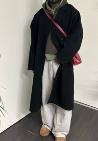 Personne portant un long manteau noir par-dessus plusieurs sweat-shirts à capuche superposés, un pantalon de survêtement gris clair, des chaussons camel et un sac bandoulière rouge, debout à l'intérieur sur un sol sombre.