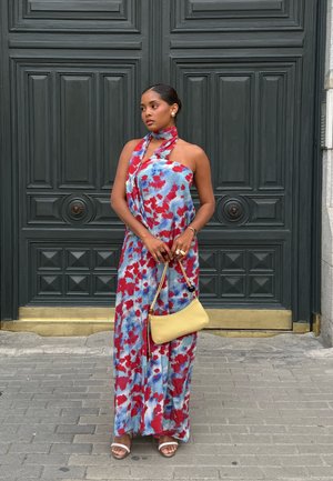 Vestido maxi con estampado floral en rojo y azul sobre tela azul claro. Escote halter anudado, complementado con un bolso amarillo y sandalias blancas.