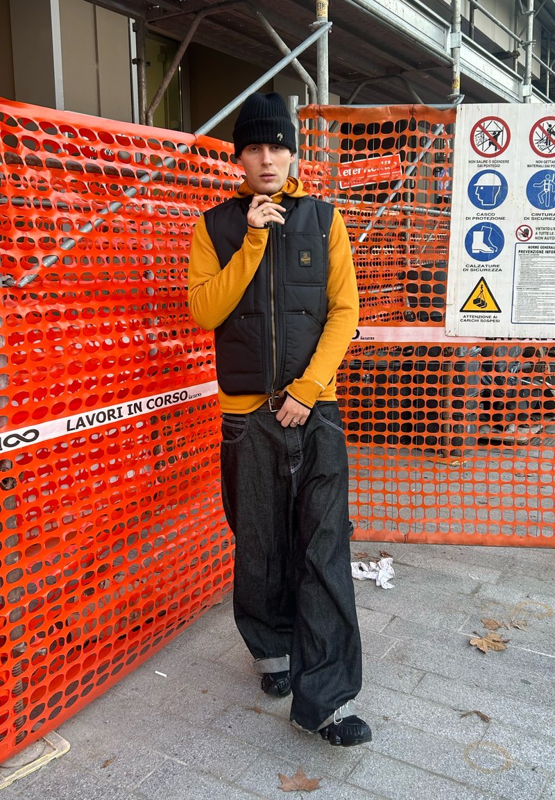 Gilet noir sans manches sur une chemise orange à manches longues, associé à un jean ample foncé et des chaussures noires, devant une clôture de sécurité orange.
