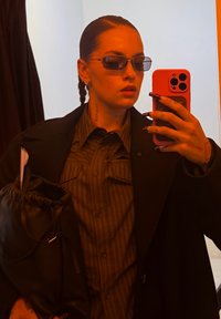 Manteau noir par-dessus une chemise rayée marron, tenant un sac à cordon noir. Porte des lunettes de soleil foncées, avec une coiffure tressée et de longs ongles.