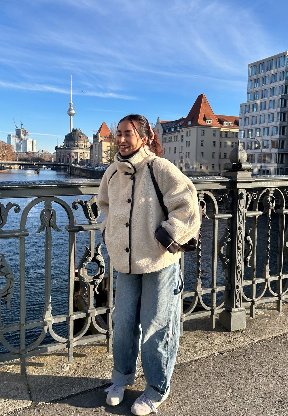 Young woman in a white fleece jacket stands smiling on a bridge over a river with historic and modern buildings in the background.